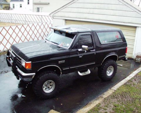 1989 Ford Bronco - A lifted black 1989 Ford Bronco sits in a wet driveway, reflecting the overcast sky.