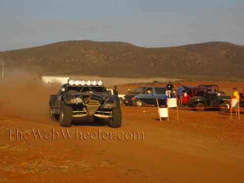 1990 Ford Bronco - long-travel off-road buggy with a roof light bar races down a dusty red-dirt course past parked pit trucks and onlookers.