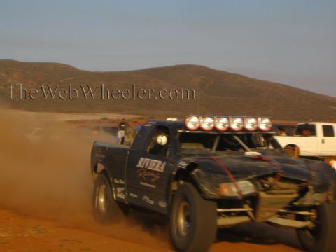 1990 Ford Bronco - dark off-road race truck with a roof-mounted light bar charges through a cloud of dust on a desert dirt course.