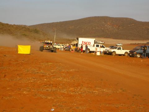 1990 Ford Bronco - desert off-road race pit area with a TSCO box truck, parked pickups and a yellow course-marker panel beside the red dirt track.