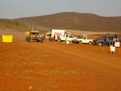 1990 Ford Bronco - off-road buggy pulls away from a pit area lined with a TSCO truck, pickups and spectators on a red dirt course at dusk.