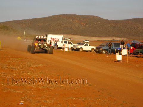 1990 Ford Bronco - off-road race buggy travels along a red dirt course past a TSCO box truck and pit vehicles, with desert hills behind.