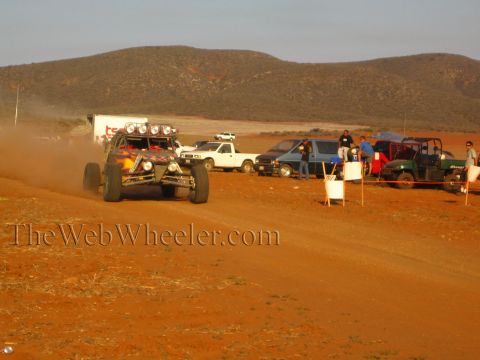 1990 Ford Bronco - open-frame off-road buggy kicks up dust as it races a red dirt course past parked pickups and spectators in the desert.
