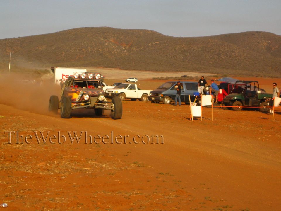 Open-frame off-road buggy kicking up dust on dirt course past parked trucks