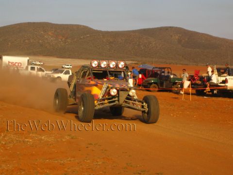 1990 Ford Bronco - flame-painted open-frame off-road buggy with a roof light bar races a red dirt course past pit vehicles and spectators.