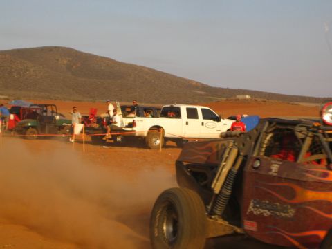 1990 Ford Bronco - white crew-cab pickup sits in a desert pit area as a flame-painted off-road buggy passes through dust in the foreground.