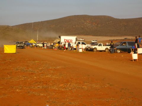 1990 Ford Bronco - wide view of a desert off-road race pit lined with a TSCO box truck, pickups, a tent and spectators along the red dirt track.