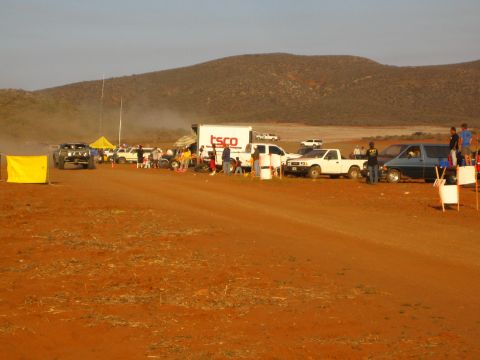 1990 Ford Bronco - row of pit vehicles and a TSCO box truck line the red dirt track at a dusty desert off-road race.