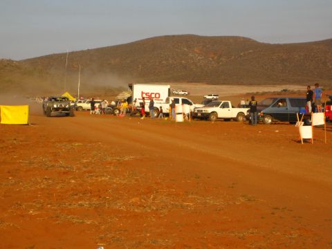 1990 Ford Bronco - off-road buggy raises dust beside a TSCO box truck and a line of pit vehicles along a red dirt desert course.