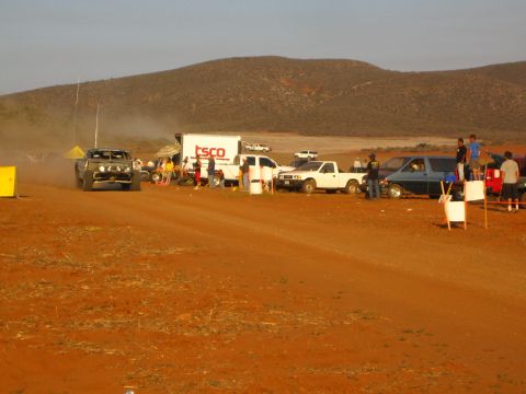 1990 Ford Bronco - off-road buggy travels a red dirt course past a TSCO box truck and pickups, with low desert hills in the background.