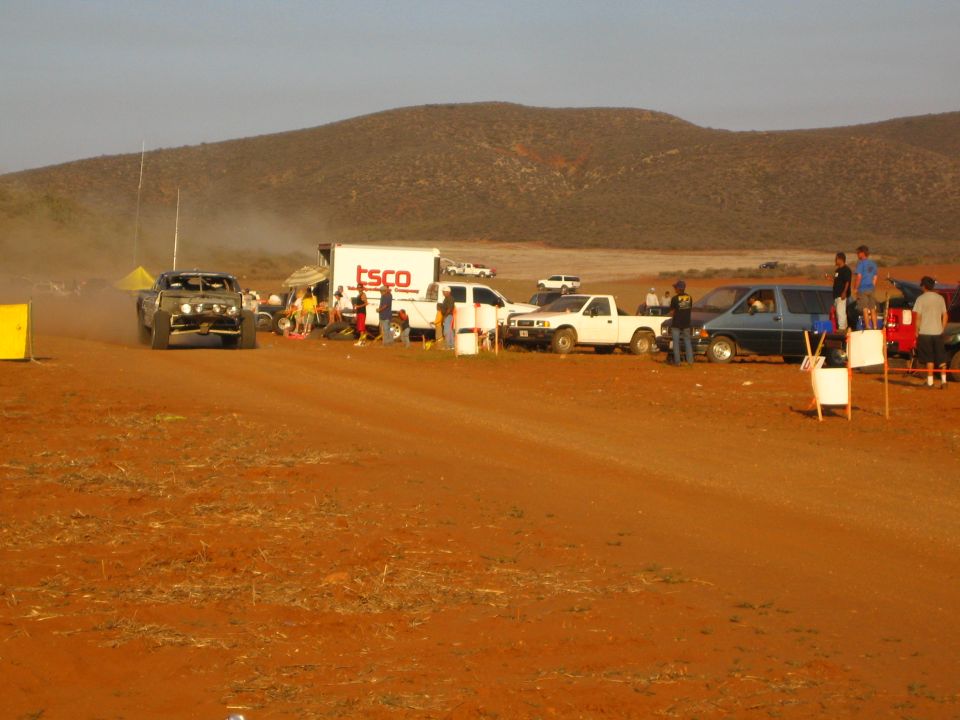 Off-road buggy on red dirt course past TSCO truck and pickups under desert hills