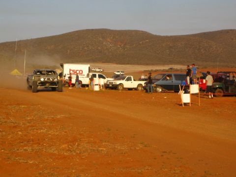 1990 Ford Bronco - off-road buggy idles in dust beside a TSCO box truck and parked pit vehicles on a red dirt course in golden light.