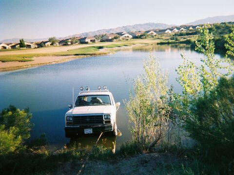 1983 Ford Bronco - A white SUV is parked partially in a body of water, with houses and hills in the background.