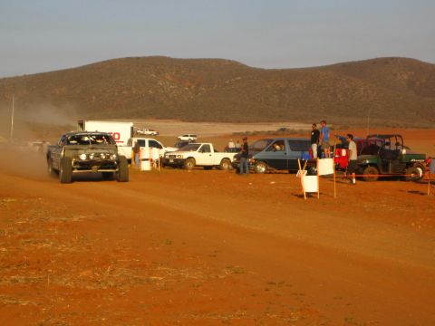 1990 Ford Bronco - off-road race truck travels a red dirt course past a TSCO box truck, pickups and a side-by-side at a desert event.