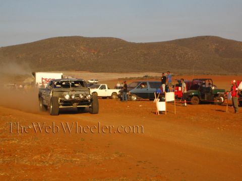 1990 Ford Bronco - dark off-road race truck corners on a red dirt course past spectators and parked pit vehicles in the desert.
