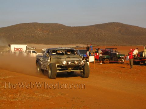1990 Ford Bronco - off-road race truck with lit headlights speeds down a red dirt course past pit vehicles and spectators in the desert.