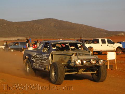 1990 Ford Bronco - blue off-road race truck marked Pro Comp and number 5 runs a red dirt course past parked pickups in the desert.