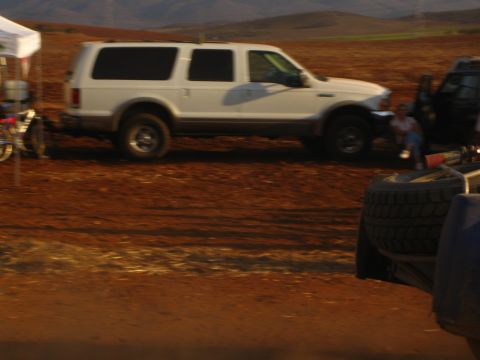 1990 Ford Bronco - white full-size SUV is parked on muddy red dirt beside a tent and an off-road race vehicle's tire and frame.