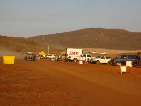 1990 Ford Bronco - desert off-road race pit area with a TSCO box truck, pickups, tents and spectators stretched along the red dirt track.