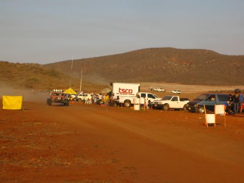 1990 Ford Bronco - off-road buggy raises dust beside a TSCO box truck and a row of pit vehicles on a red dirt course under desert hills.