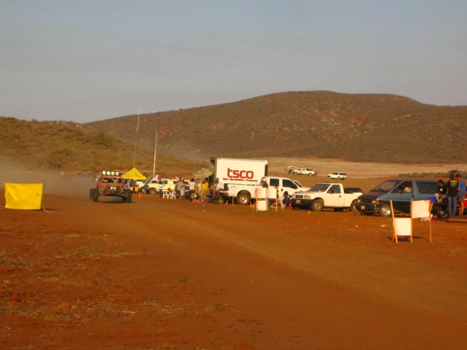 Buggy in dust beside TSCO truck and pit vehicles on red dirt under hills