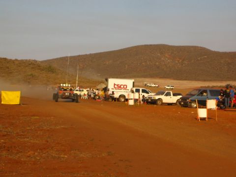 1990 Ford Bronco - off-road buggy with a roof light bar passes pit vehicles and a TSCO box truck along a red dirt desert course.