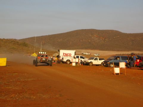1990 Ford Bronco - red off-road buggy travels a red dirt course past a TSCO box truck and pickups, with low desert hills behind.