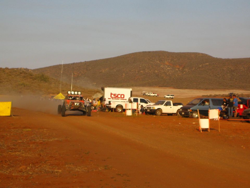 Red off-road buggy on dirt course past TSCO truck and pickups under desert hills