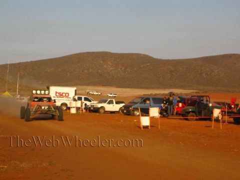 1990 Ford Bronco - orange tube-frame off-road race buggy speeds along a dusty desert road past a row of parked spectator trucks and a TSCO box trailer at suns…