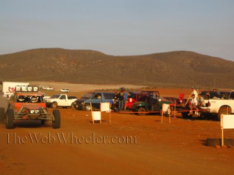 1990 Ford Bronco - orange off-road race buggy rolls along a dirt road with a long line of parked pickups, a trailer and spectators behind it in golden desert…