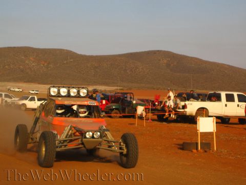 1990 Ford Bronco - off-road race buggy marked 1009, fitted with a roof light bar, drives down a dusty desert road past parked trucks and a UTV at sunset.
