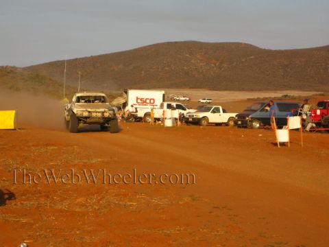 1990 Ford Bronco - tan off-road race truck kicks up dust on a desert road while a TSCO box trailer and several pickups sit parked at the pit area in evening l…