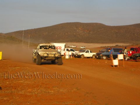 1990 Ford Bronco - silver tube-frame off-road race truck speeds along a dirt desert road past parked pickups and a box trailer in warm sunset light.