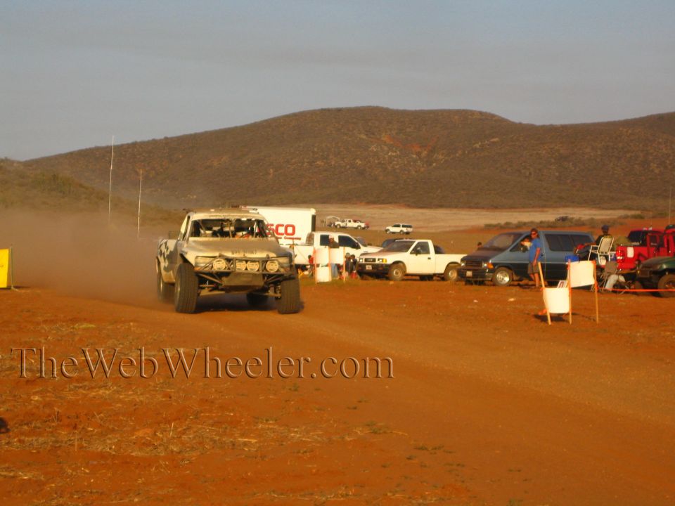 Silver off-road race truck on dirt road past pit trucks and trailer at sunset