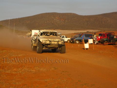 1990 Ford Bronco - tan trophy-truck-style off-road racer drives toward the camera on a dusty desert road with parked vehicles and spectators along the pit.