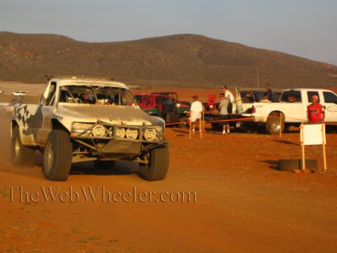 1990 Ford Bronco - silver off-road race truck with a roof light bar travels a dirt road as spectators and pickups line the desert pit area at sunset.