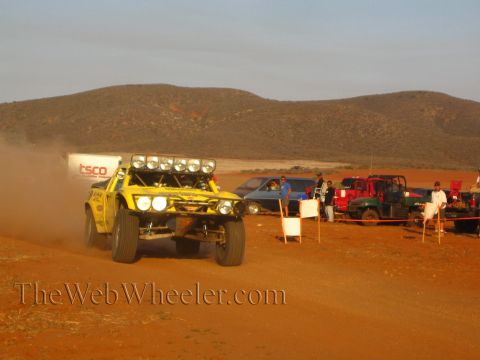 1990 Ford Bronco - yellow tube-frame off-road race truck with a roof light bar speeds down a dirt road past a TSCO trailer and parked vehicles at sunset.