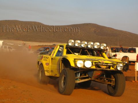 1990 Ford Bronco - yellow off-road race truck with a six-lamp roof light bar charges through desert dust past spectators in low golden light.