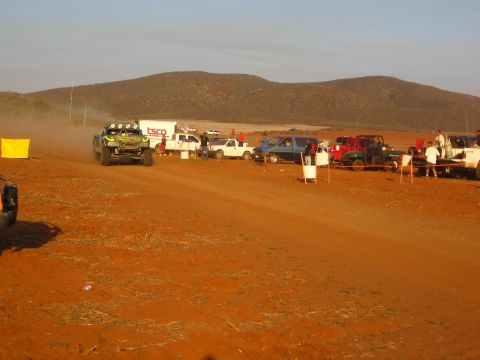 1990 Ford Bronco - distant off-road racer drives along a dirt road as a long line of pickups, trailers and spectators fills the desert pit area at sunset.
