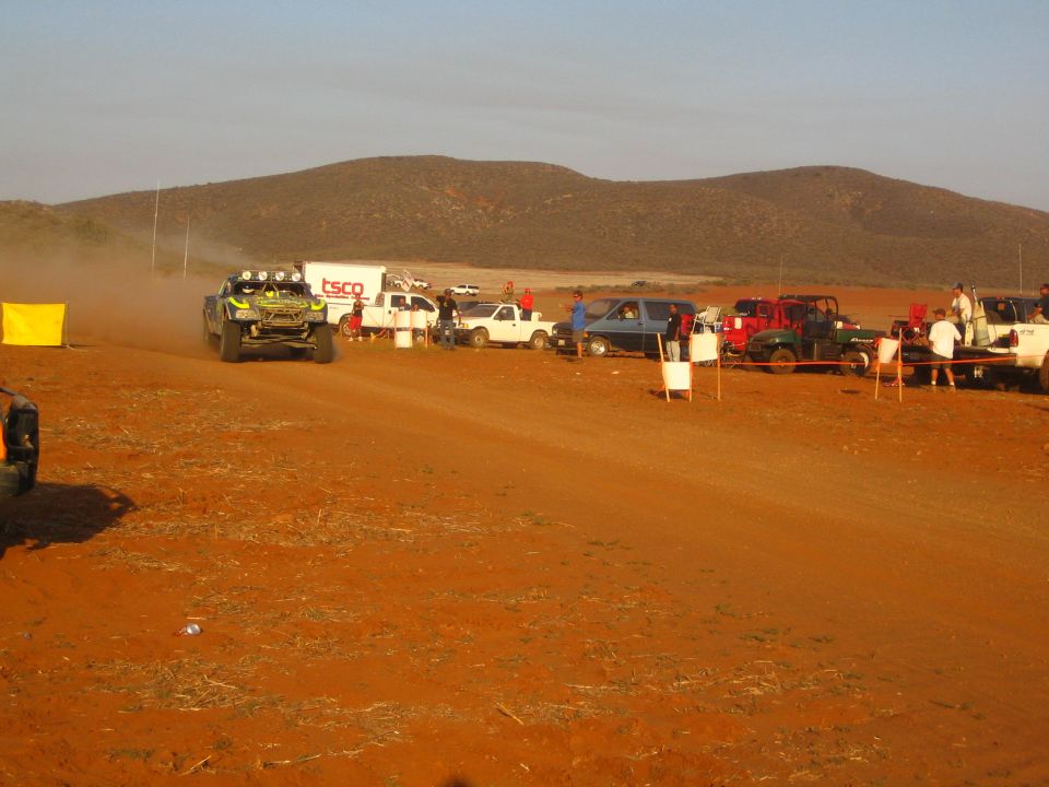 Off-road racer on distant dirt road past long line of pit vehicles in desert