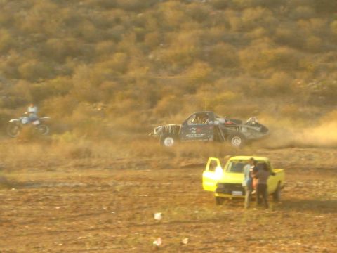 1990 Ford Bronco - off-road race truck catches air over scrubby desert terrain as a dirt bike rider and a yellow pickup with people look on.