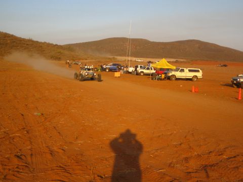 1990 Ford Bronco - off-road buggy drives a red-dirt course past a yellow pit canopy and parked trucks, with the photographer's shadow cast across the foregrou…