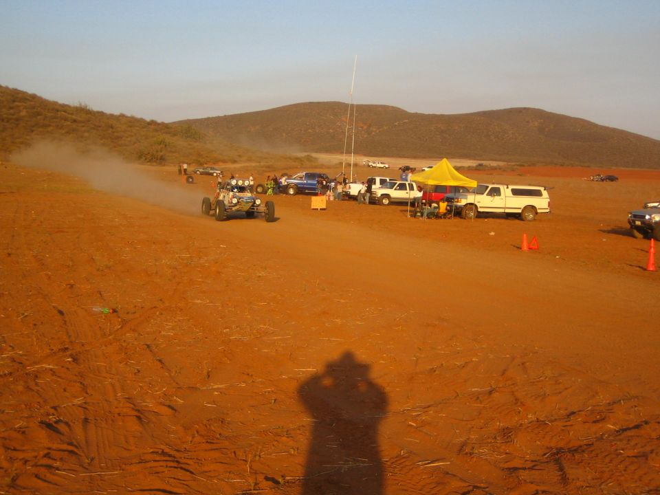 Off-road buggy on red dirt course past pit canopy and trucks, photographer shadow