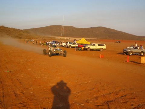 1990 Ford Bronco - tube-frame off-road buggy travels a red-dirt road past a yellow pit canopy and parked pickups, the photographer's shadow in the foreground.