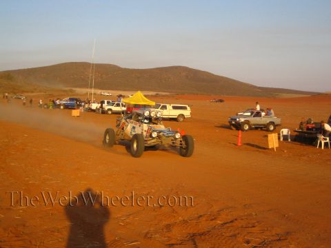 1990 Ford Bronco - off-road buggy with a roof light cluster drives a red-dirt road past a yellow pit canopy, parked pickups and seated spectators at sunset.