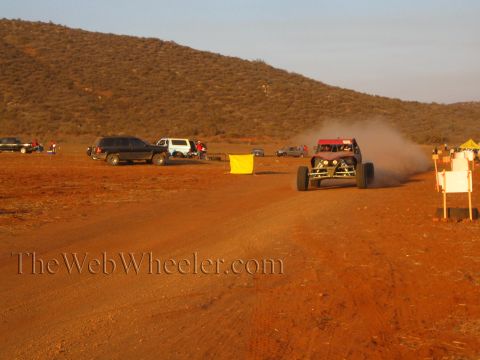 1990 Ford Bronco - off-road buggy raises dust on a desert dirt road past parked SUVs, a yellow course flag and white course-marker signs at sunset.