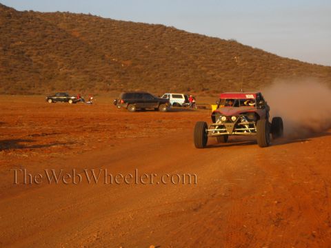 1990 Ford Bronco - red off-road buggy marked 108 speeds down a red-dirt desert road, trailing dust past parked cars and SUVs in evening light.
