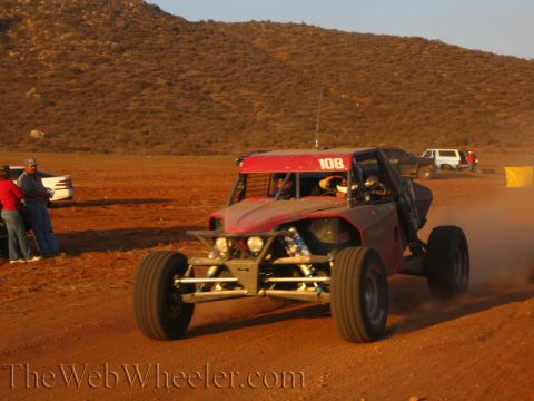 1990 Ford Bronco - red tube-frame off-road buggy numbered 108 corners on desert dirt with its rear suspension exposed as spectators watch from the roadside.