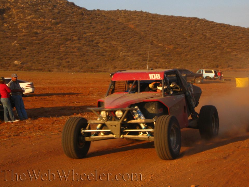 Red number 108 tube-frame off-road buggy cornering on dirt as spectators watch