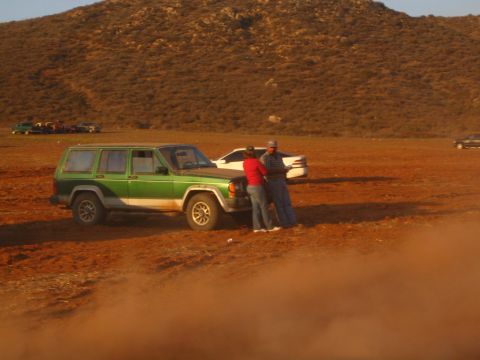 1990 Ford Bronco - Two men stand talking beside a green and tan Jeep Cherokee parked on red dirt in an open desert area with a brushy hillside behind.